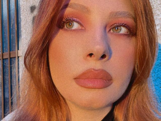 MiraVolt has red hair, pink eyeshadow, and glossy pink lips. Her green eyes look slightly to the side. She has a fair skin tone and is wearing black eyeliner.