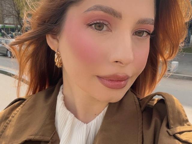 MiraVolt has pink blush on her cheeks, brown wavy hair, and is wearing a brown jacket with gold earrings. Her pink lipstick and brown eyes are visible.