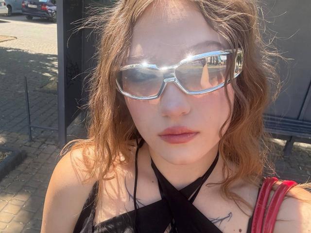 Bubochka, con i capelli castano ondulati, indossa occhiali argentati lucidi, un top nero con spalline sottili e una cinghia rossa. Ha labbra rosa chiaro e piccoli tatuaggi sul petto. Il sole si riflette sugli occhiali..