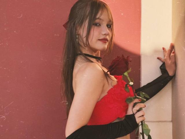 Bubochka, con lunghi capelli castani, un reggiseno rosso, un collare nero e maniche nere, tiene una rosa e si trova di fronte a un muro rosso e bianco. Il suo seno è visibile..