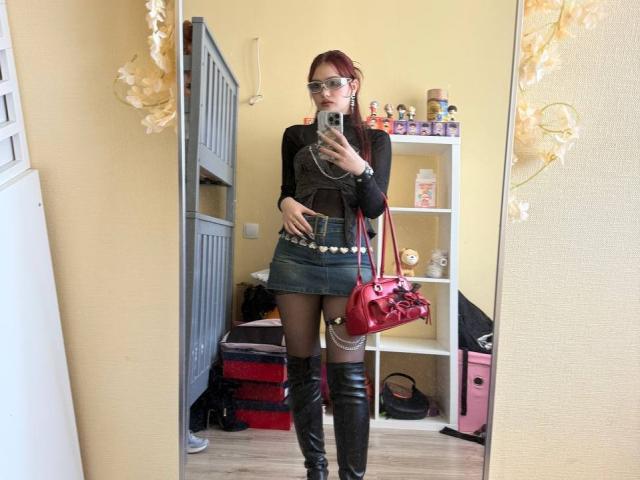 Bubochka with red hair, black top, green mini-skirt, black thigh-high boots, red purse, holding phone, mirror selfie, yellow walls, gray cabinet, white shelves, colorful toys.