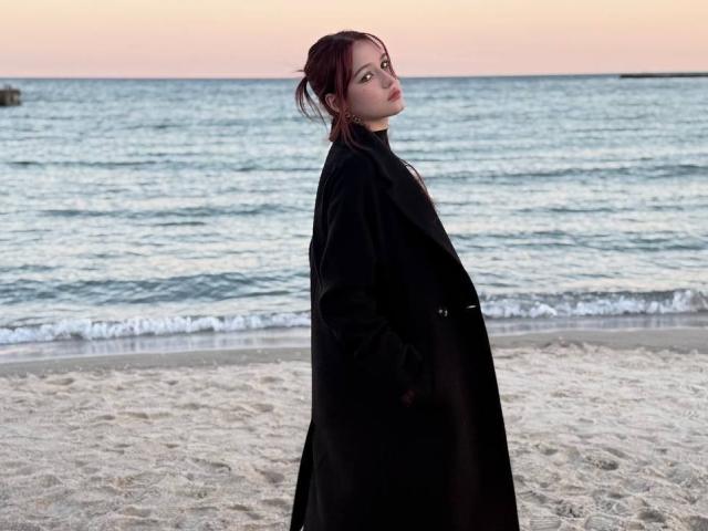 Bubochka stands on a beach, wearing a long black coat, with hands in pockets, looking back over shoulder, her red hair tied up, ocean and sunset in background.