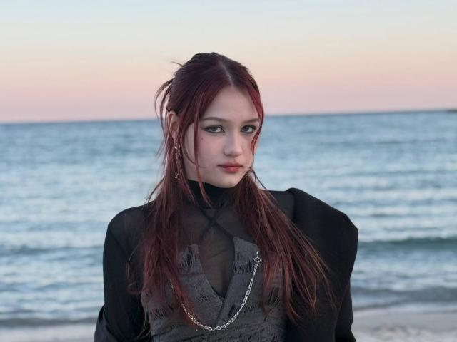 Bubochka with red hair, pale skin, black sheer top, chain necklace, looking at camera, standing by the ocean at sunset, slight smirk, small nose, dark eyeliner, subtle makeup.