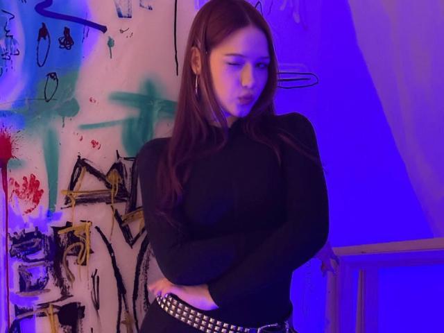 Bubochka with brown hair, black long-sleeve top, crossed arms, studded belt, blue-purple light, looking down, standing, wooden railing on right.