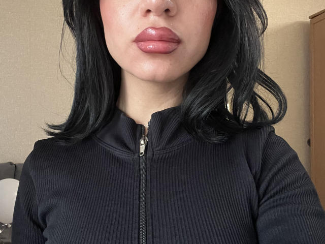 CharleenOachs with glossy pink lips, black wavy hair, and a black rib-knit zip-up shirt, showing her upper body and slightly parted zipper. Her skin is fair.