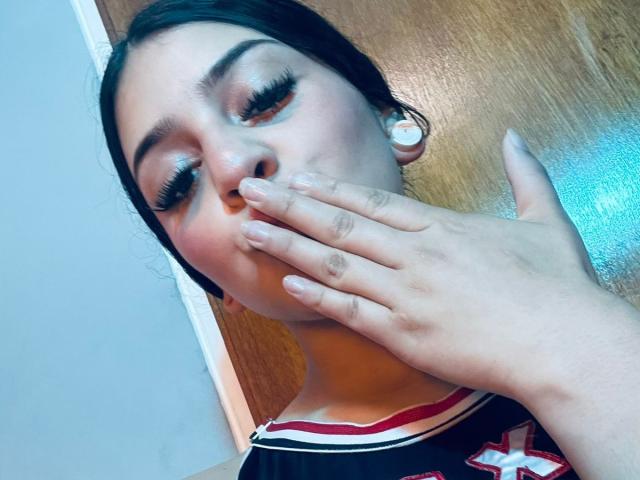LonxEva with long eyelashes, fair skin, black hair, and nude nails, covering her mouth with her hand, wearing a black top with red and white trim.