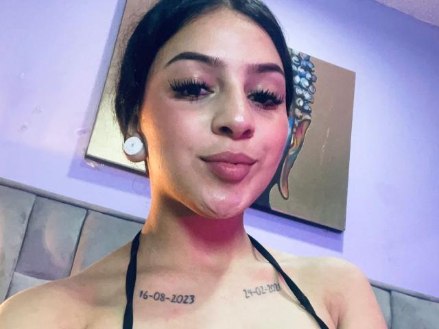 LonxEva has dark hair, thick eyebrows, long eyelashes, and a slight smile. She's wearing a black halter top, showing her tits, and has white earbuds in. Her face is glistening.
