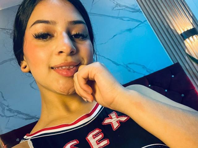 LonxEva with medium skin tone, dark hair, and long eyelashes, smiling, touching her chin, wearing a black and red cheerleader top, showing her pouty lips and subtle makeup.