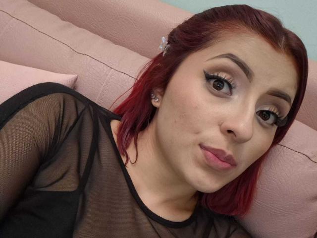 LizBonnet with red hair, wearing a sheer black top, has pouty lips, eyeliner, and a nose piercing, looking at the camera with a slightly puckered expression.