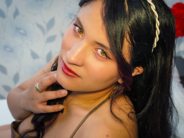 AlisonXDuarte ha gli occhi marroni, il rossetto rosso e i capelli scuri con le punte viola, indossa una fascia dorata e una collana. Si sta toccando il mento, mostrando un anello d'oro. Il seno è visibile..