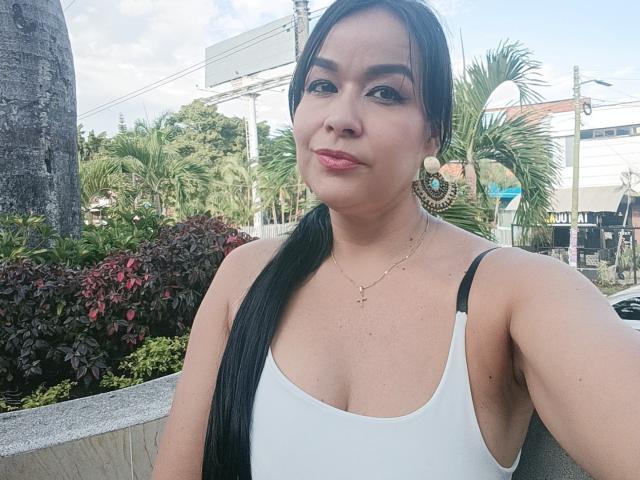 TamaraFoxy with long black ponytail, white tank top, showing small tits, pink lipstick, gold earrings, and necklace, outside, clear sky, palm trees, and urban background in photo.