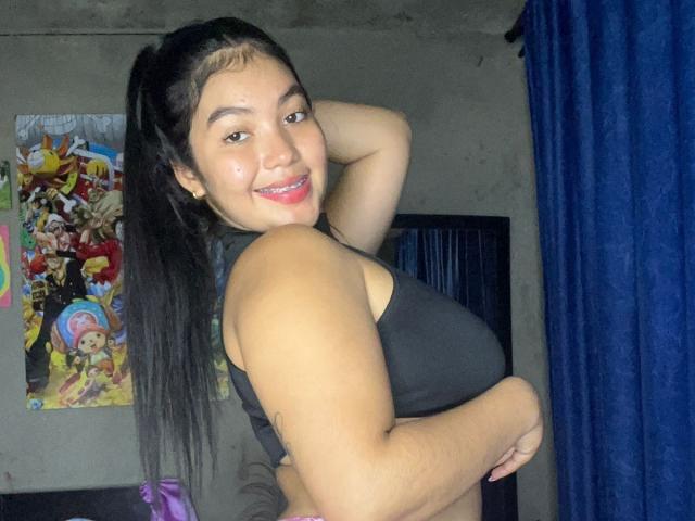 AlisBella with long black hair, smiling, wearing a black sports bra, showing her tits, hand behind head, blue curtain, anime poster, pink bag, and concrete wall in background.