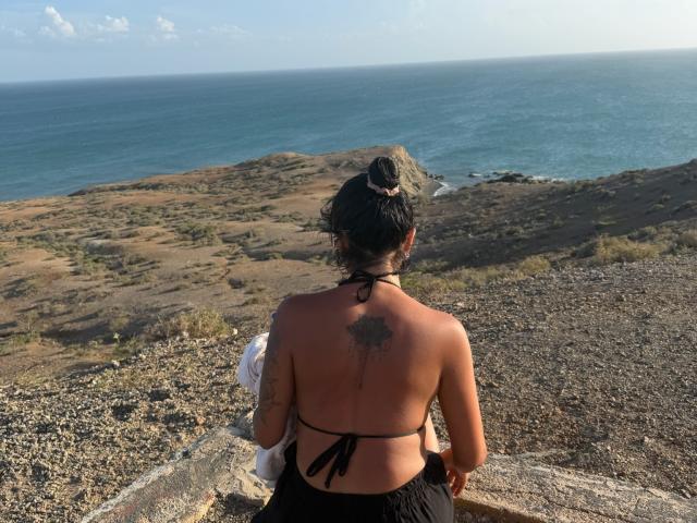MajoJones with a black halter top, showing her bare back and a tattoo, stands on a rocky cliff, facing the ocean, her dark hair in a bun.