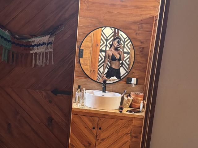 MajoJones taking a mirror selfie, showing her tits in a black bra, standing in a wooden bathroom with a round mirror, wood countertop, and toiletries.