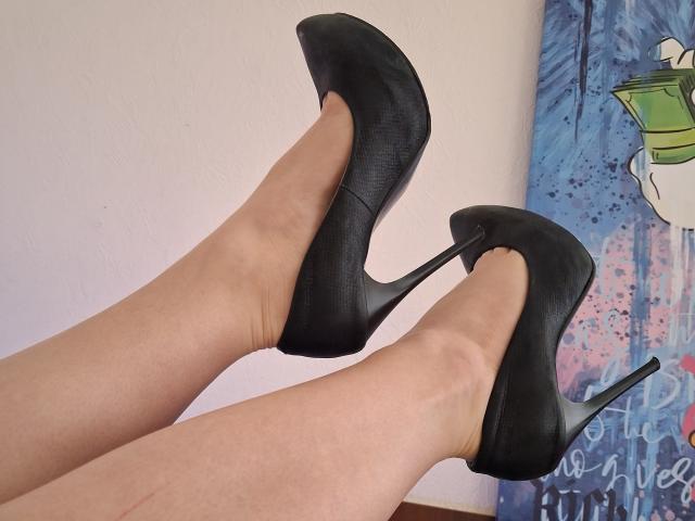 Marriam's legs in black stiletto heels, showing off her high heels with pointed toes and thick heels. Her legs are fair-skinned, and the heels are sleek and shiny.