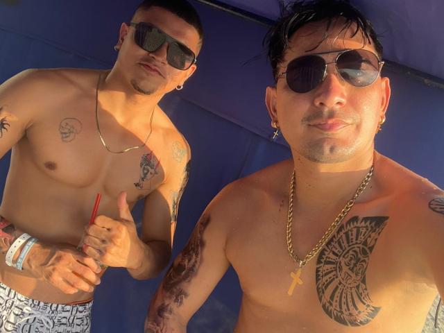 Two shirtless, tattooed WilFelixes with dark hair, sunglasses, and gold chains. WilFelix on left holds red straw, giving thumbs-up. WilFelix on right has large chest tattoo.