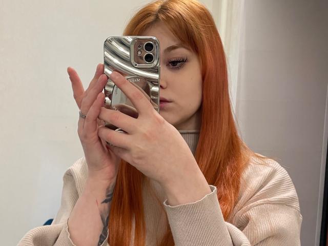 LettyRussum, met lang oranje haar, houdt een zilveren telefoon met een golvend patroon vast en maakt een selfie in een spiegel. Ze draagt een beige geribbelde coltrui en toont een tatoeage op haar hand..