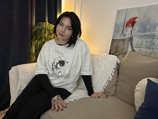BueEyesOcean mit schwarzen Haaren, einem weißen T-Shirt, schwarzen Ärmeln und schwarzen Hosen sitzt auf einem beigen Sofa, blickt zur Seite und lächelt leicht, ihre Hände liegen auf ihrem Oberschenkel..