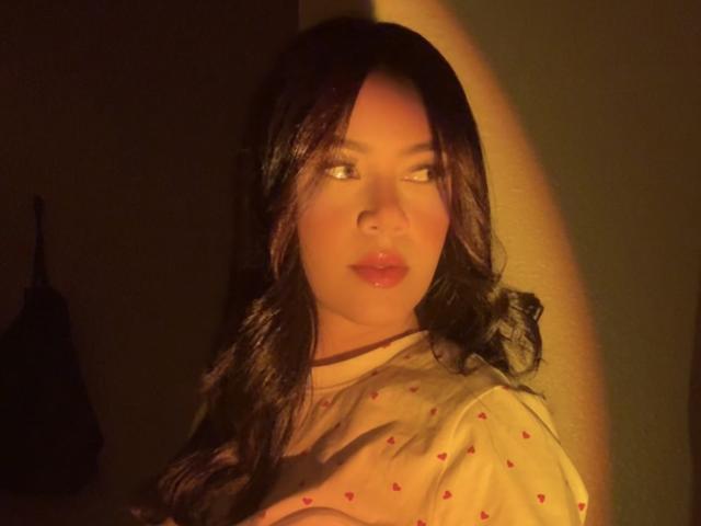 Yuliannyw, avec ses cheveux noirs ondulés, une lumière orange éclatante, regardant de côté, portant un haut transparent avec des cœurs rouges, paraissant mignonne avec des lèvres brillantes, fond sombre..
