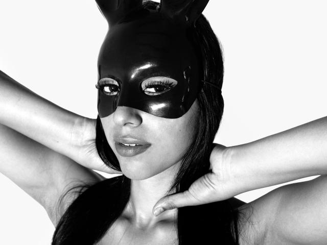 SiennaLanne with long dark hair, wearing black bunny ears and a mask, covering her mouth with her finger, showing her bare shoulders and a tattoo on her arm. Black and white.