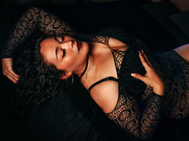 ArianaCherry with curly black hair, eyes closed, wearing a see-through black mesh dress, red lipstick, and a black choker, lying on a black quilt, hand on chest.