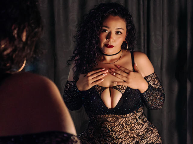 ArianaCherry, con cabello negro rizado, lápiz labial oscuro y aros grandes, vestida con un vestido negro de encaje con un escote pronunciado, con las manos sobre los senos, en una habitación con poca luz..