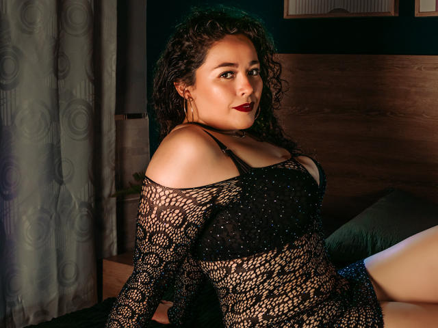 ArianaCherry mit lockigem, braunem Haar, dunkelrotem Lippenstift, trägt ein schwarzes, durchsichtiges, Netzoberteil, das ihre Brüste und ihr schwarzes BH-Top freilegt, sitzt auf einem Bett und schaut in die Kamera..