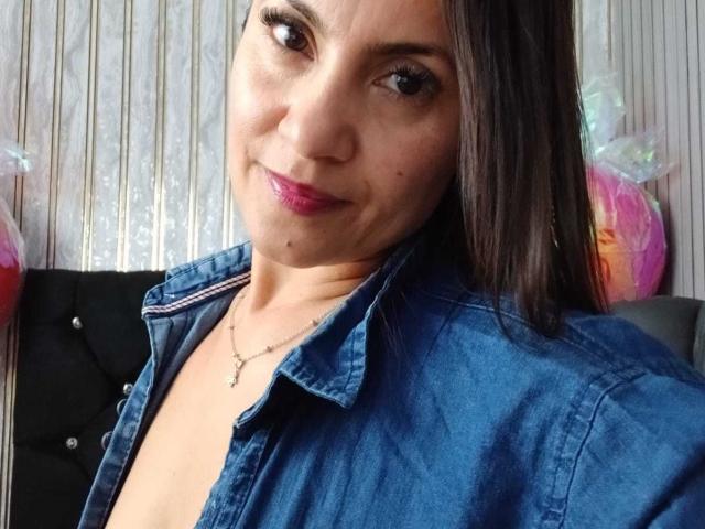SolaraDubois with medium tits, wearing an open blue denim shirt, has pink lipstick, brown eyes, and long dark hair, smiling slightly, necklace with small pendant.