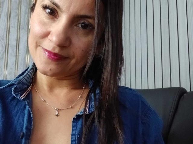 SolaraDubois with dark brown hair, pink lipstick, denim shirt, small necklace with cross, smiling at camera, visible moles on face, black leather chair behind her.