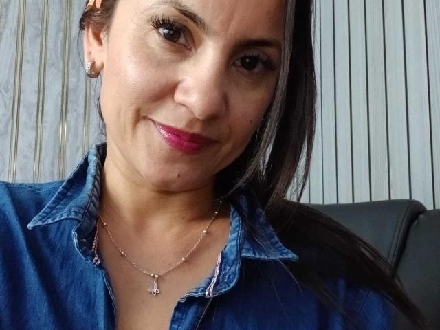 SolaraDubois with pink lipstick, dark straight hair, wearing a blue denim shirt, silver necklace with a cross, and small earrings, smiling slightly. Her tits are modestly visible.