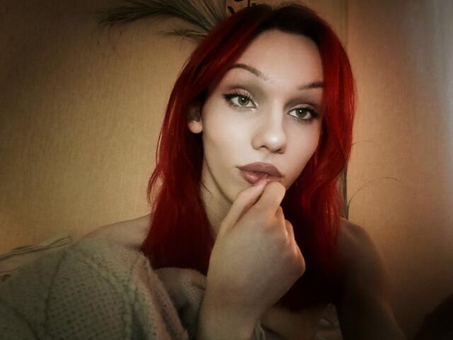 Red-haired KiranaHayes with green eyes, pouty lips, and finger touching her mouth, wearing a grey sweater, looking directly at the camera with a slightly serious expression.