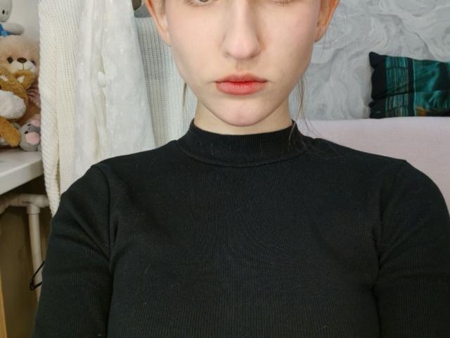 KrystaFessel mit heller Haut, roten Lippen und braunen Haaren, trägt einen schwarzen, gerippten Rollkragenpullover und blickt mit neutralem Gesichtsausdruck direkt in die Kamera. Weicher, weißer Hintergrund..