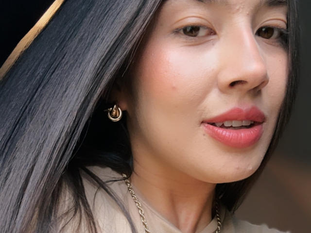 EmmaLombardi's face, showing smooth brown skin, straight black hair, gold earrings, pink lipstick, and a subtle chain necklace, with a slight, cute smile.