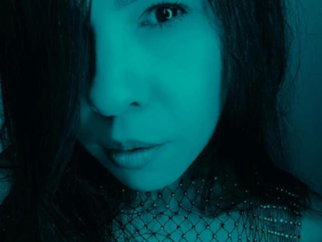 Le visage d'AlinaOwens, avec une peau teintée de bleu, des cheveux ondulés foncés qui recouvrent un œil, portant un collier de perles, les lèvres légèrement entrouvertes et un sourire discret. Son œil est visible..