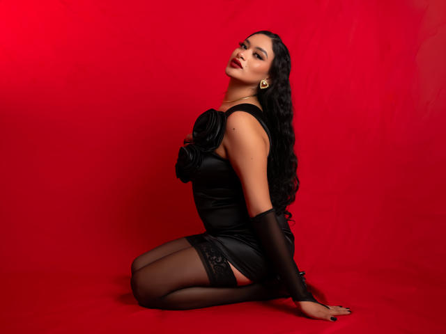 Adalina Vera, arrodillada, con un vestido de satén negro, medias hasta el muslo, y cabello negro largo y rizado, fondo rojo, y expresión sensual, mostrando su trasero y sus labios rojos brillantes..