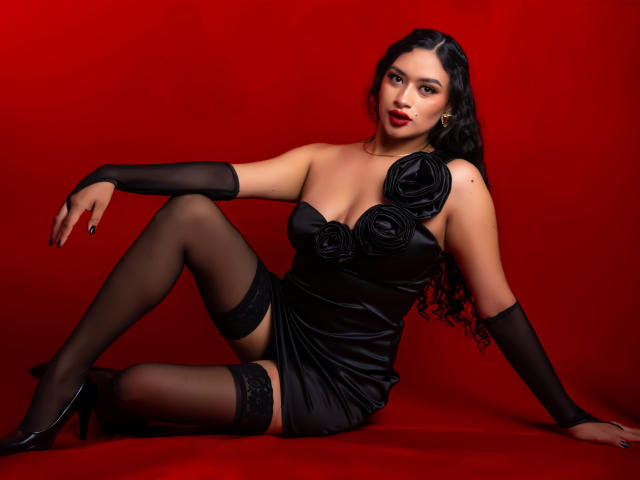 Adalina Vera con vestido negro, adornos de rosas, guantes negros, medias hasta el muslo y tacones negros, sentada sobre un fondo rojo, con las piernas cruzadas, mirando a la cámara, con cabello negro largo y rizado, y lápiz labial rojo..