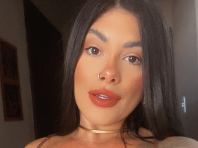 FerrazJuanitta69, con cabello negro largo, ojos marrones, lápiz labial rojo, un piercing en la nariz, un collar dorado y un rostro sexy y atractivo. No se ven los senos, pero se ve muy atractiva..