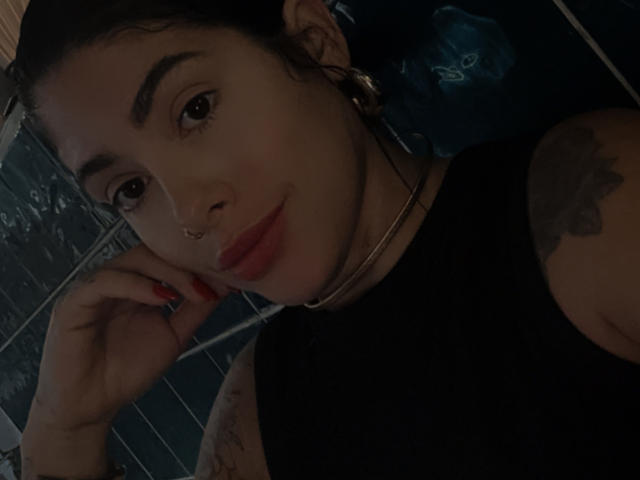 FerrazJuanitta69 mit dunklem Haar, rotem Lippenstift, Nasenring, schwarzem Top ohne Ärmel, Tattoos und roten Nägeln, liegend, in die Kamera blickend, leichtes Lächeln, dunkler, gefliester Hintergrund..