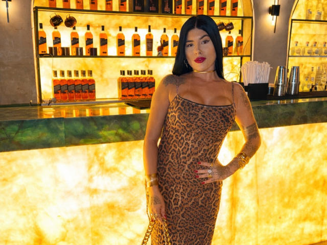 FerrazJuanitta69 stands in a bar, wearing a tight leopard print dress that highlights her tits and ass. She has tattoos, dark hair, and red lipstick.