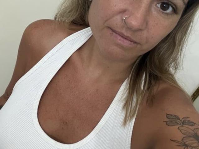 BellissimoFiore mit gebräunter Haut, Nasenring und einem leichten Lächeln, trägt ein weißes Rippstrick-Tanktop, das kleine Brüste und ein Tattoo an ihrer rechten Schulter zeigt..