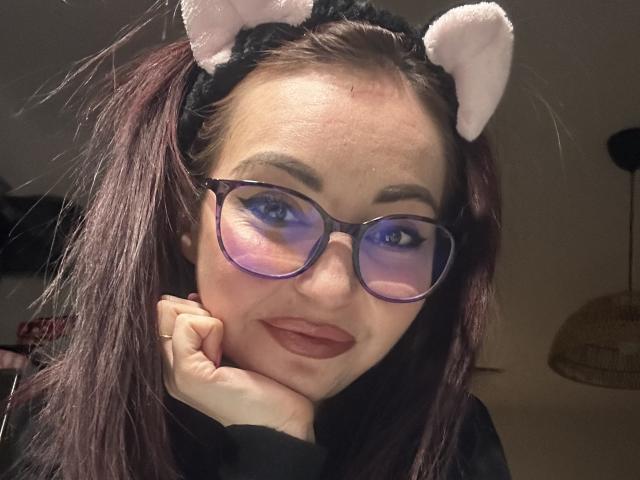 ChloeSensual with purple glasses, dark red lipstick, and pink cat ears headband, resting her chin on her hand, smiling slightly, dark brown hair, fair skin.