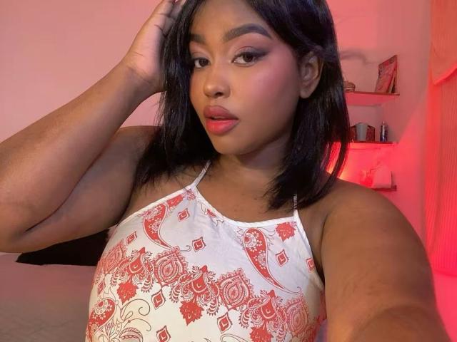 NicolZurich has dark skin, medium-length black hair, and is wearing a white halter top with red paisley patterns. Her right hand touches her head, and she has red lipstick.