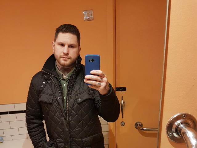 Een man met een gladde huid en een baard, gekleed in een zwarte gewatteerde jas en een groen shirt, houdt een blauwe telefoon vast en maakt een selfie in een badkamer met oranje muren. Op zijn hand zit een tatoeage..