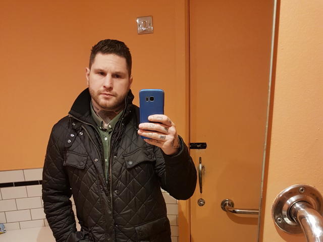 Satinsubsilky, con i capelli e la barba castani e corti, indossa una giacca nera trapuntata e una camicia verde, e si scatta un selfie allo specchio con un telefono blu in un bagno con le pareti arancioni..