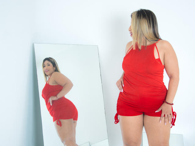 AuroraBenson stands in a red dress, looking at her reflection in a mirror. She has blonde and brown hair, medium tits, and a curvy ass. Mirror reflection.
