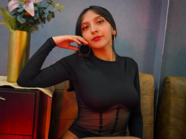 AlissierAmell, con cabello oscuro, vestida con una blusa negra transparente de manga larga que deja ver sus senos, sosteniendo un anillo negro cerca de su mejilla, sentada junto a un mueble de madera con un jarrón..