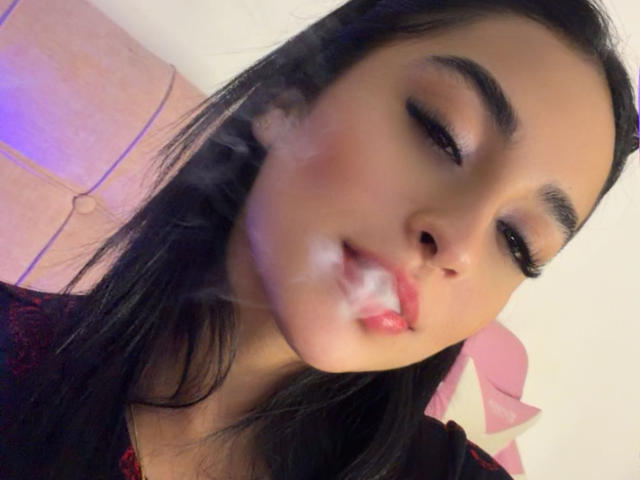 EmmaWaltter with black hair, closed eyes, and a thick white cum shoot in her mouth, wearing dark red lace. Her makeup is smoky, and her skin is tan.