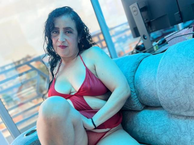 AnnitaSex69, con i capelli neri ricci, un bikini rosso e un seno di media grandezza, seduta su un divano blu, con la mano destra sulla vulva, il ginocchio sinistro sollevato, che guarda la fotocamera con un leggero sorriso..