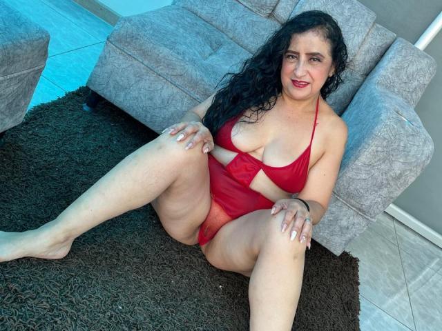 AnnitaSex69 con cabello negro largo, traje de baño entero rojo, sentada sobre una alfombra marrón, con las piernas separadas, mostrando su trasero y sus pechos, sonriendo, con las manos en las rodillas, en una sala de estar..