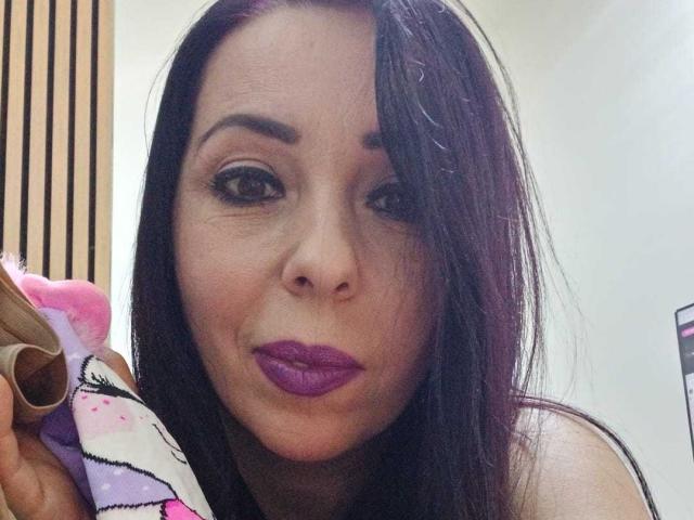 LauraSin, con cabello castaño oscuro, lápiz labial morado y sosteniendo un unicornio de peluche rosa y blanco, haciendo una mueca. Tiene ojos grandes y atractivos y una nariz linda..